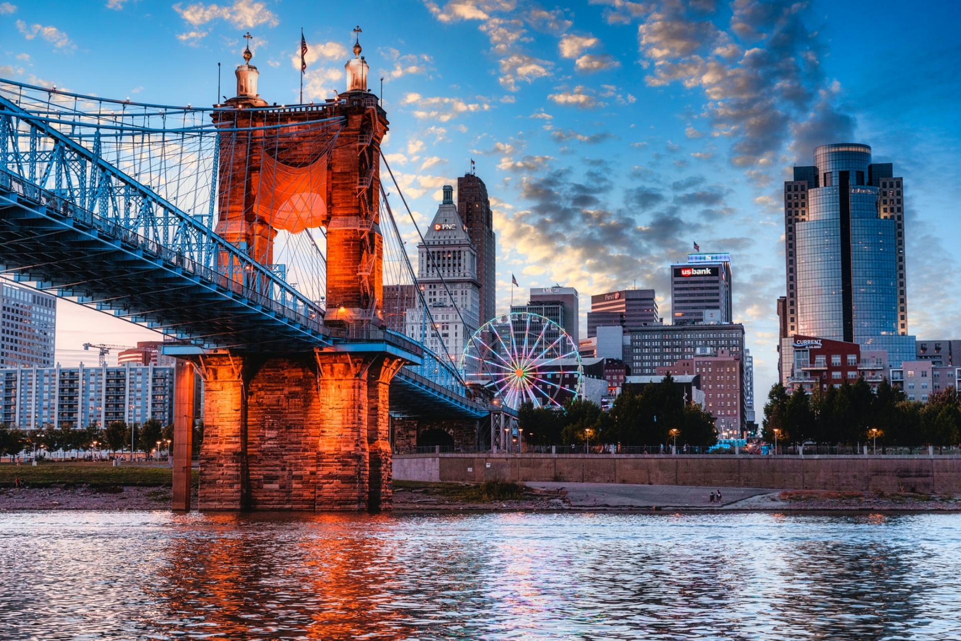  	The John A Roebling Suspension Bridge across the Ohio River and Cincinnati office buildings beneath a blue sky.