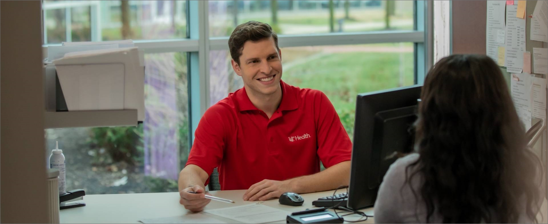 	A young male customer service rep in a red polo assists a female client at his computer station.